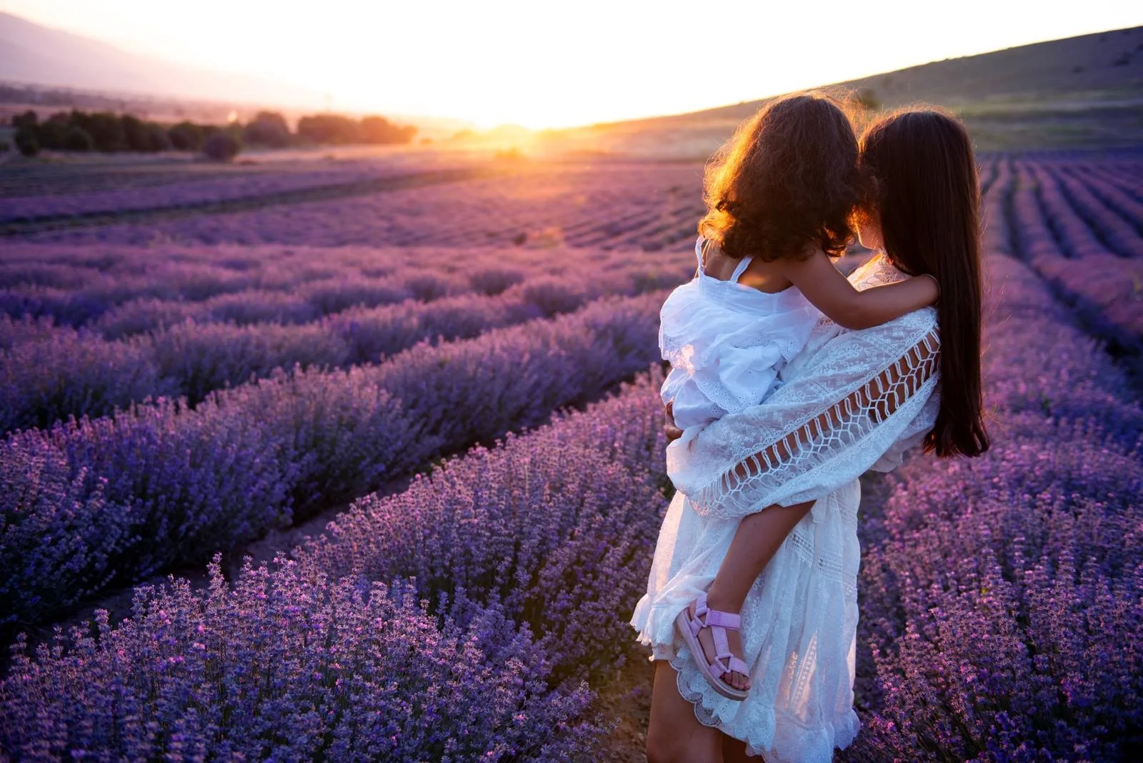 A mother carrying her daughter through a lavender field at sunset, both in white dresses, seen from behind